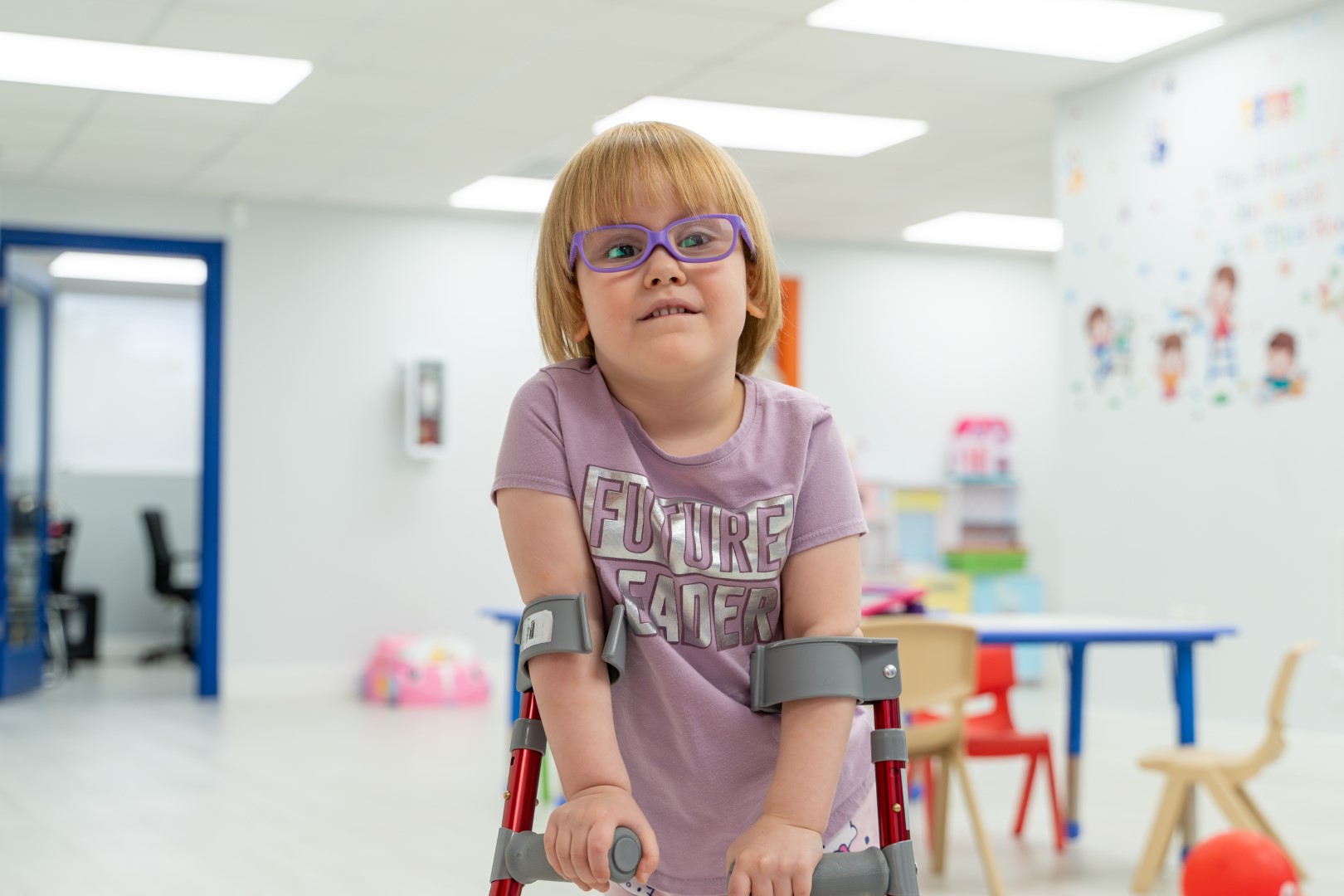 A smiling girl in glasses standing with the support of a gait trainer at Peek A Boo PPEC